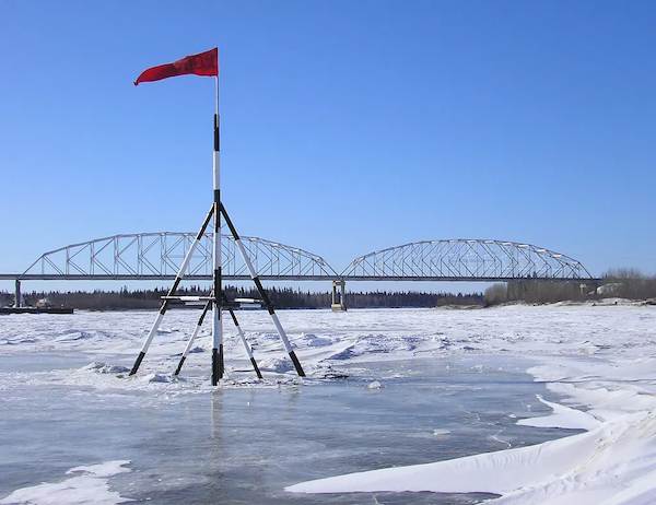 Black and white striped poles form a tripod anchored in the frozen Nenana. The tripod supports a red pennant blowing southward with the Nenana bridge in the background. Clear blue sky.